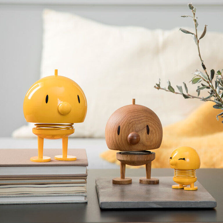 Holzfiguren der Marke Hoptimist Bumble aus Eichenholz. Drei Figuren in verschiedenen Größen und Farben, überwiegend gelb und naturfarben, auf einem Tisch in einem stilvoll eingerichteten Raum mit Büchern und Pflanzen im Hintergrund.