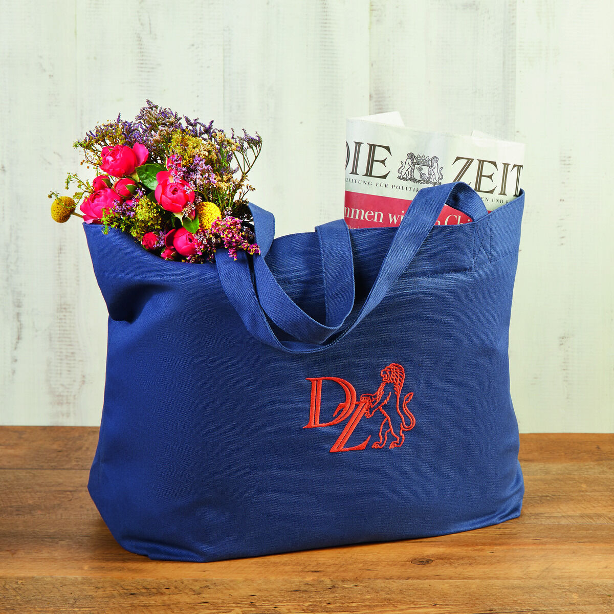 Eine blaue Canvas-Tasche mit orangenem ZEIT-Logo steht auf einem Holztisch. In der Tasche befindet sich ein bunter Blumenstrauß und eine Zeitung.