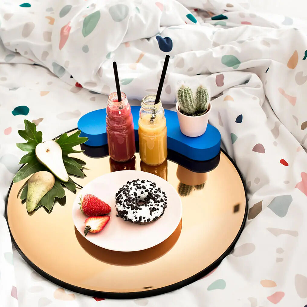 Auf einem weich fallenden, weißgrundigen Bettwäsche-Set mit buntem Terrazzo-Muster liegt ein runder, glänzender Kupfertablett. Darauf stehen zwei Gläser mit Strohhalmen, ein Donut, Obst, ein Kaktus im Topf und eine abstrakte blaue Ablage.