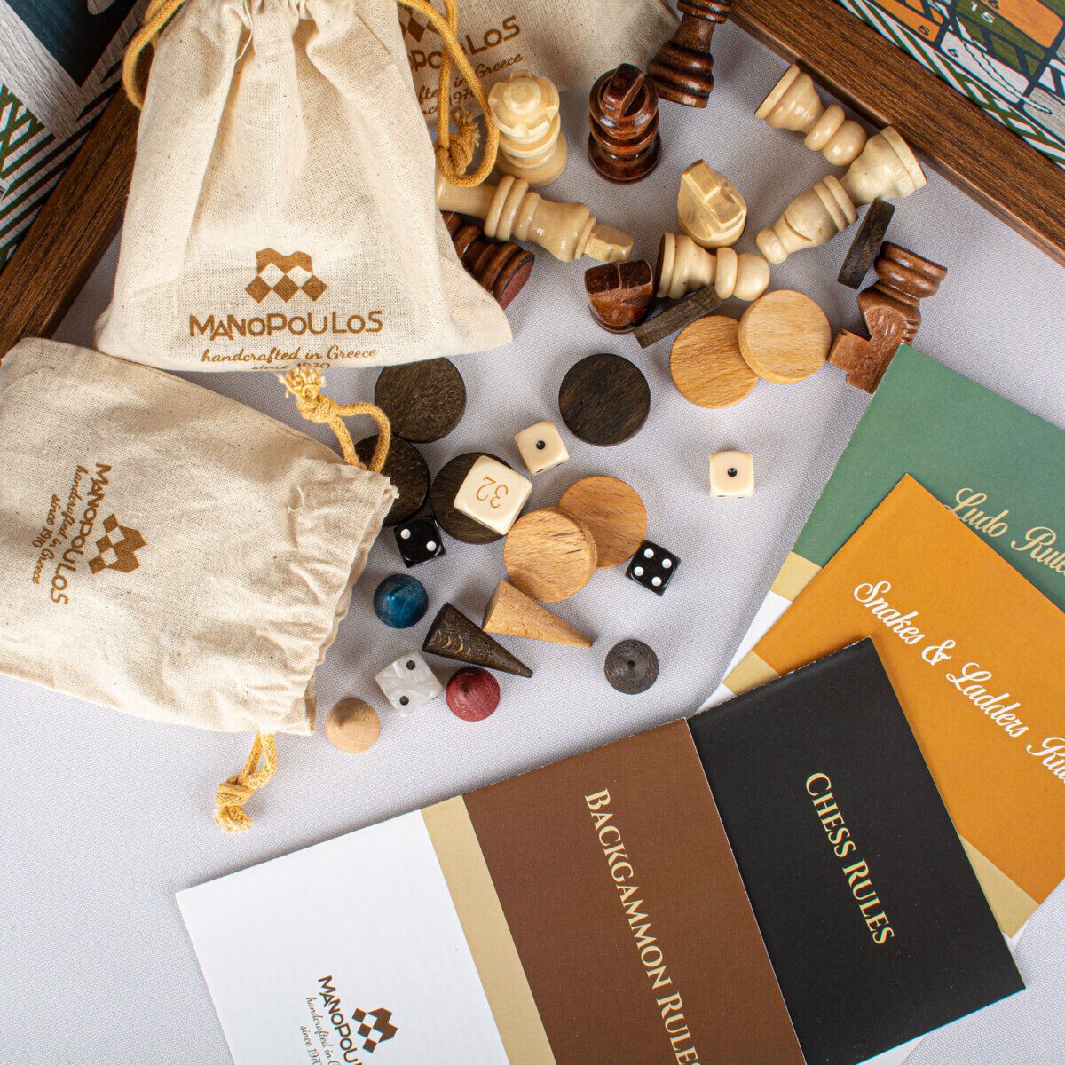Eine Sammlung von Holzspielzeugen aus Walnuss-Holz mit Schach, Backgammon, Mühlespiel und Dame Spiel. Spielsteine und Würfel sind sichtbar, sowie Spielbretter und Anleitungshefte. Die hochwertigen Taschen tragen das Logo von Manopoulos.