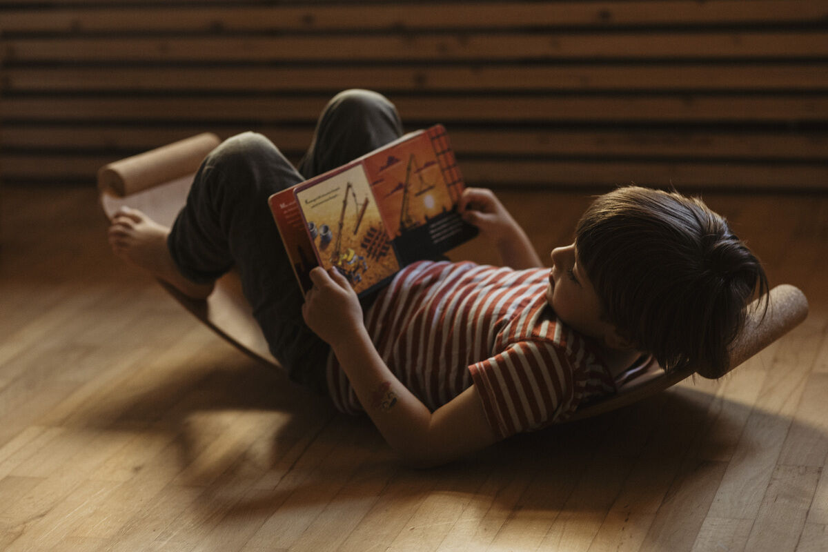 Ein Kind liegt auf einem Balance Board aus Holz und liest ein Buch. Das Holz des Balance Boards hat eine geölte Oberfläche. Die Umgebung ist in einem warmen, natürlichen Licht gehalten.