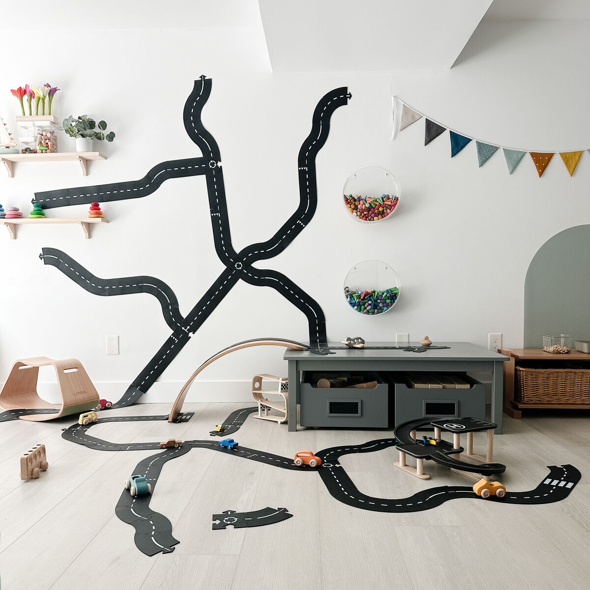 Ein Kinderzimmer mit einem aufgebauten Straßenbau-Set »King of the road« auf dem hellen Fußboden. Das Set besteht aus schwarzen, flexibel aneinandergelegten Straßenteilen und kleinen Holzautos. Im Hintergrund sind eine Kommode, Wanddekorationen und diverses Kinderspielzeug zu sehen.