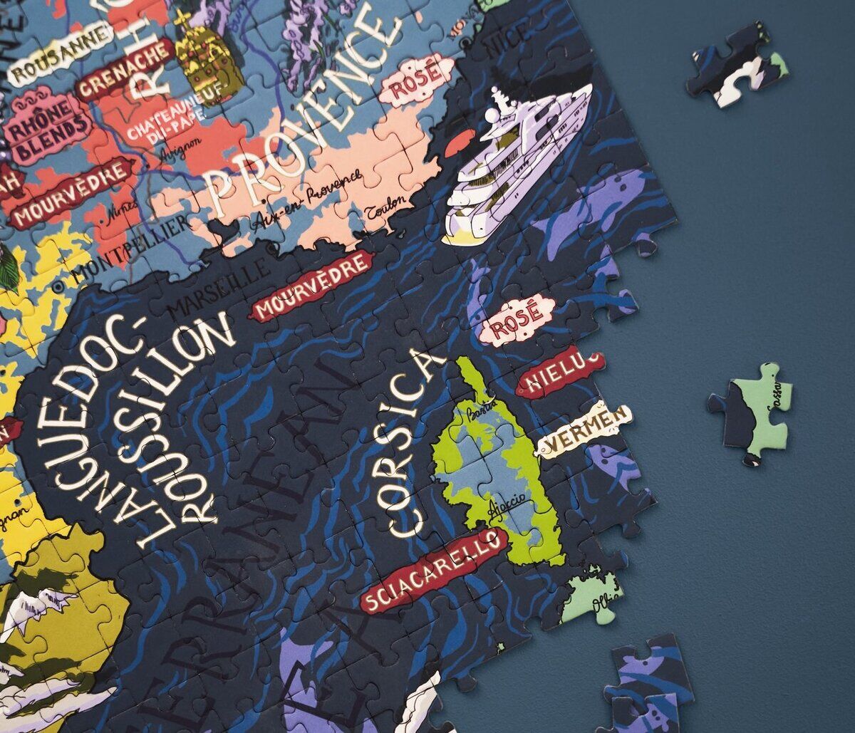Puzzle mit einer detaillierten Karte von Frankreichs Weinregionen, die auf einem dunkelblauen Hintergrund liegt. Einige Puzzleteile sind bereits zusammengesetzt, während andere Teile daneben liegen. Die Karte zeigt mehrere bekannte Weinregionen wie Languedoc, Roussillon und Provence, sowie kleinere Bereiche und Gewässer, die die geografischen Merkmale hervorheben.