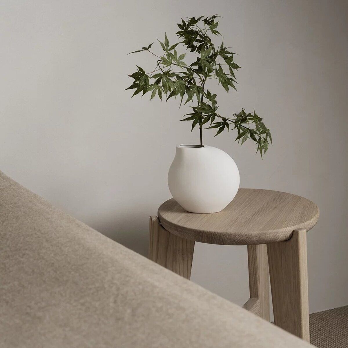 Ein minimalistischer Sitzhocker aus hellem Holz steht vor einer neutralen Wand. Auf dem Hocker steht eine weiße, runde Vase mit einem grünen Zweig. Das Bild vermittelt eine ruhige, moderne Wohnatmosphäre.