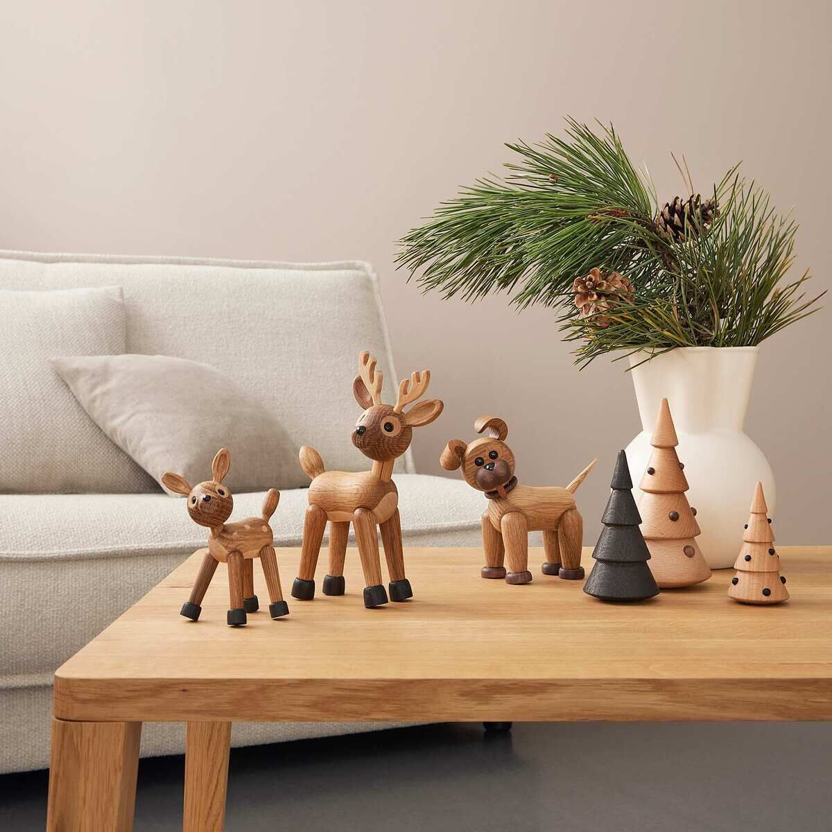 Mehrere Holzfiguren in Tierform, darunter ein Rentier und ein Bär, stehen zusammen mit dekorativen Holzbäumchen auf einem Holztisch in einem modern eingerichteten Wohnzimmer. Im Hintergrund steht eine weiße Vase mit Tannenzweigen auf einem hellen Sofa.