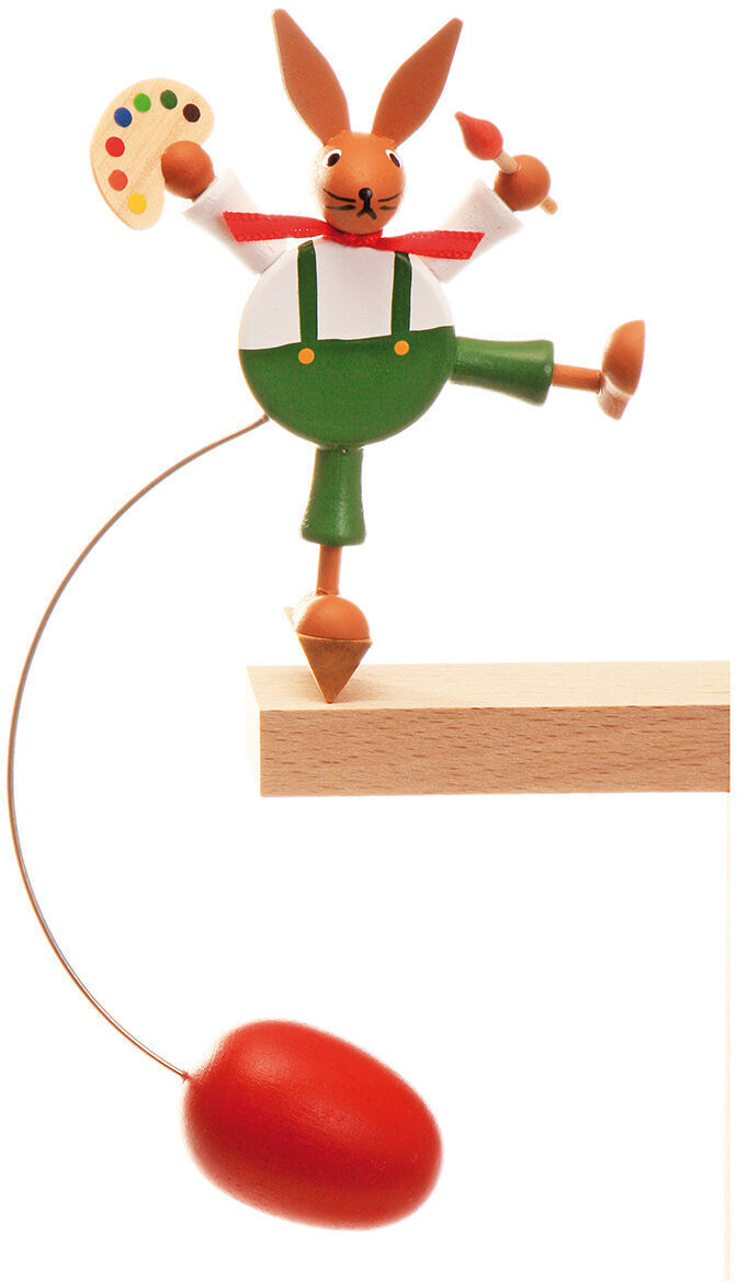 Holzfigur eines Osterhasen mit roter Hose und grüner Uniform, der eine Farbpalette hält und auf einem kleinen Holzständer balanciert. Unten hängt ein rotes Ei an einem Draht.