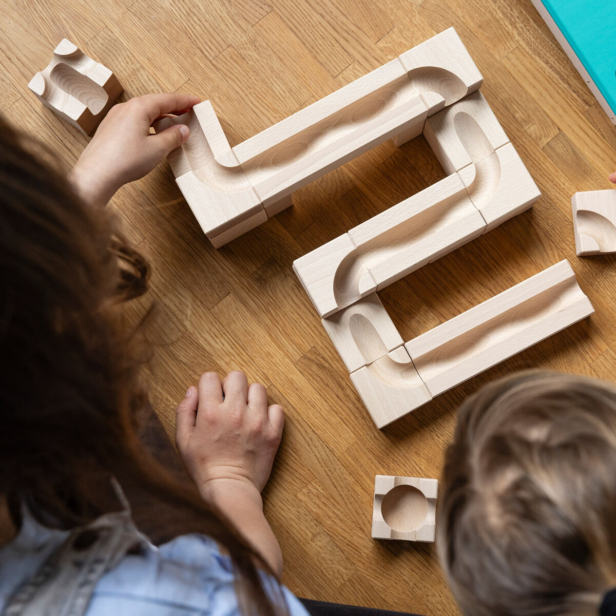 Holz-Kugelbahn auf einem Holztisch, von Kinderhänden zusammengesteckt. Eine Box in Blau liegt daneben. Verschiedene Teile der Kugelbahn sind sichtbar.