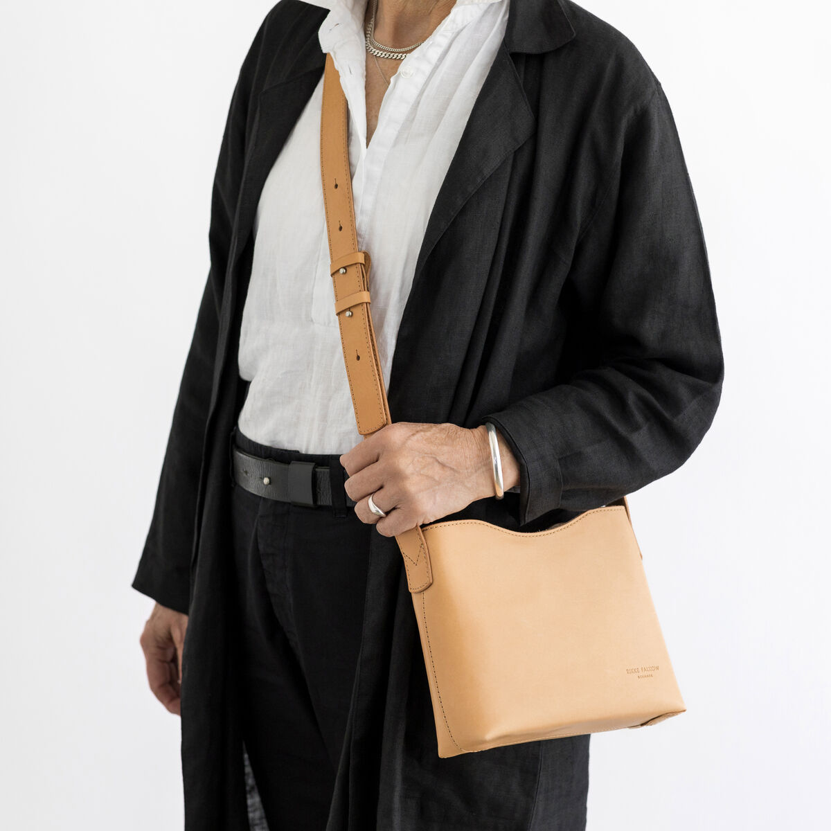 Eine Person trägt eine beige Schultertasche aus Kernleder über einer schwarzen Jacke und einem weißen Hemd. Die Tasche hat einen minimalistischen und eleganten Stil, perfekt für den Alltag oder besondere Anlässe.