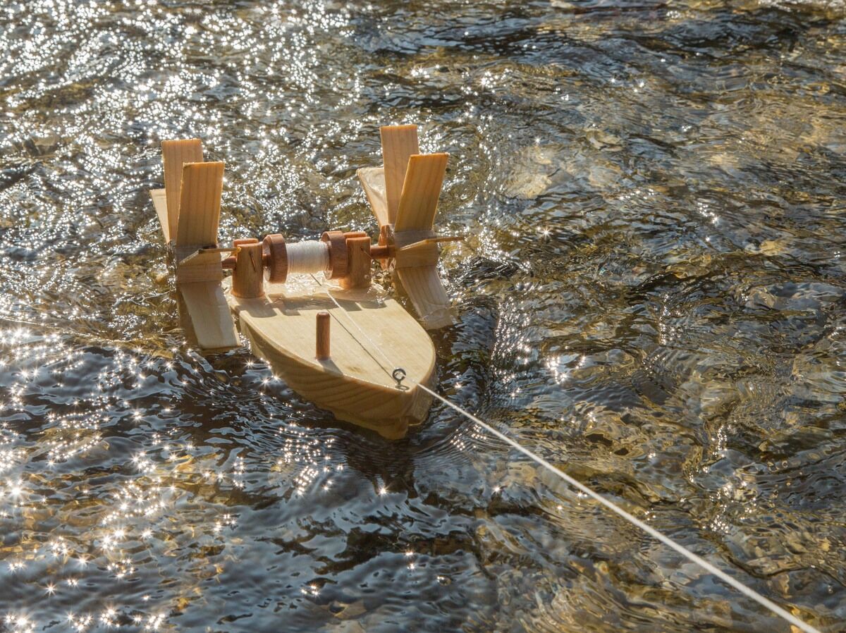 Ein Holzboot aus einem Bausatz 'Forelle' schwimmt auf einem Fluss. Das Wasser glänzt und reflektiert das Sonnenlicht. Das Boot ist mit einer Schnur festgemacht.