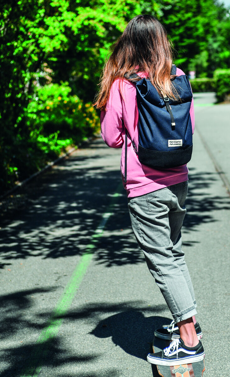 Frau mit langem, mittelbraunem Haar in rosa Kapuzenpullover und grauer Hose fährt auf einem Skateboard einen grünen Fahrradweg entlang und trägt einen dunkelblauen Rucksack namens 'Fahrradtasche Annecy'. Im Hintergrund sind Bäume und eine Straße zu sehen.