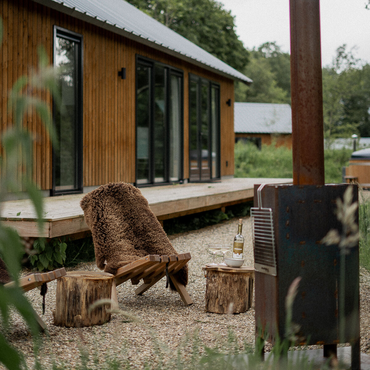 Ein braunes Schafsfell mit Lederschlaufe liegt auf einer Holzbank im Außenbereich vor einem modernen Holzhaus. Daneben stehen rustikale Holzstühle und ein Metallofen, umgeben von grüner Natur, was eine gemütliche, naturnahe Atmosphäre vermittelt.