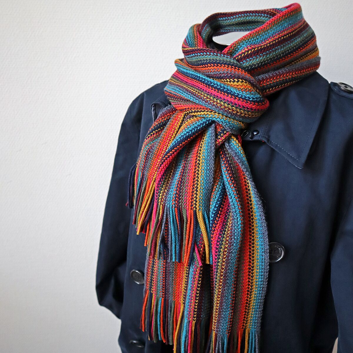 Ein gestrickter Merinoschal in den Farben Rot und Blau mit weiteren bunten Streifen liegt dekorativ um die Schultern eines dunkelblauen Mantels. Der Schal wirkt weich, warm und stilvoll – ideal für kalte Tage.
