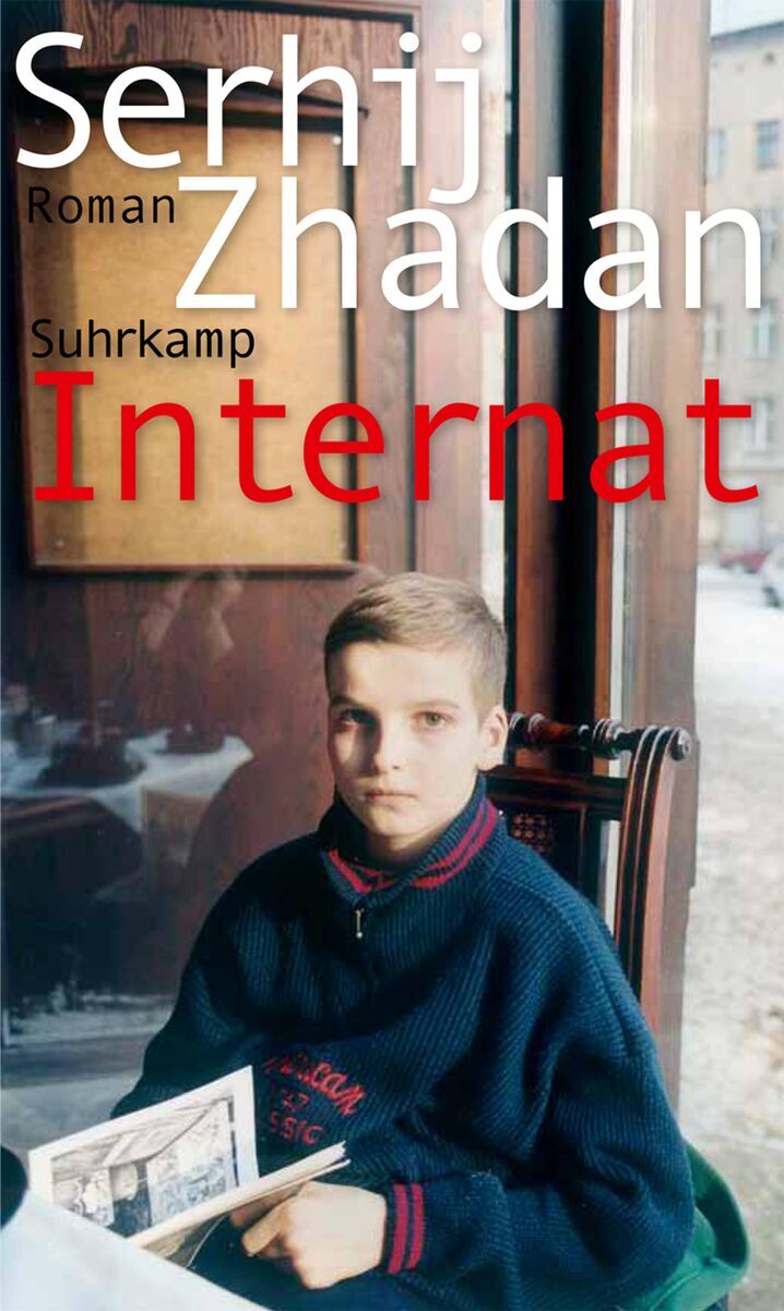 Das Cover des Buches 'Internat' von Serhij Zhadan zeigt einen jungen Jungen in einem blauen Pullover mit einem Buch in den Händen. Im Hintergrund sind ein Fenster und ein verschwommenes Gebäude zu sehen. Der Titel des Buches sowie der Name des Autors und der Verlag sind in Weiß und Rot geschrieben.