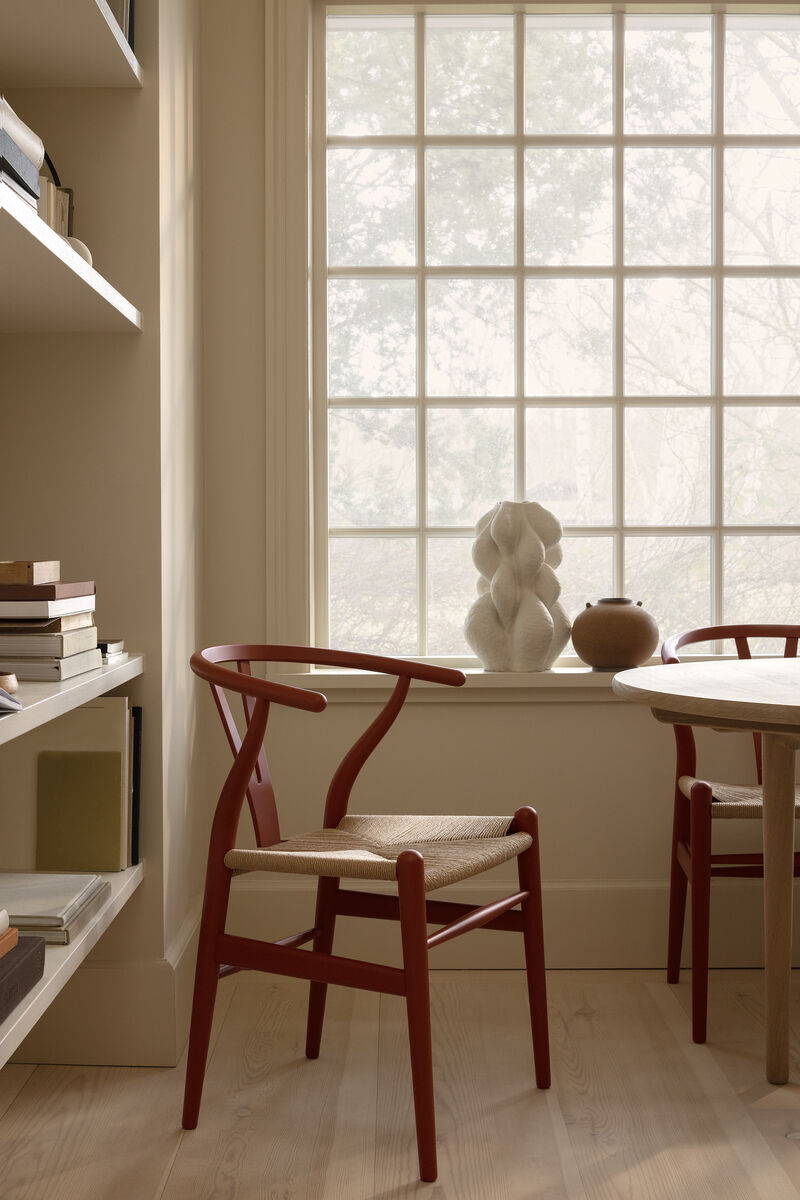 Ein moderner, roter Stuhl »Stuhl CH24 Soft« von Carl Hansen steht in einem minimalistisch eingerichteten Raum neben einem runden Tisch und einem Fenster. Im Hintergrund befindet sich ein Bücherregal und auf der Fensterbank stehen dekorative Objekte.