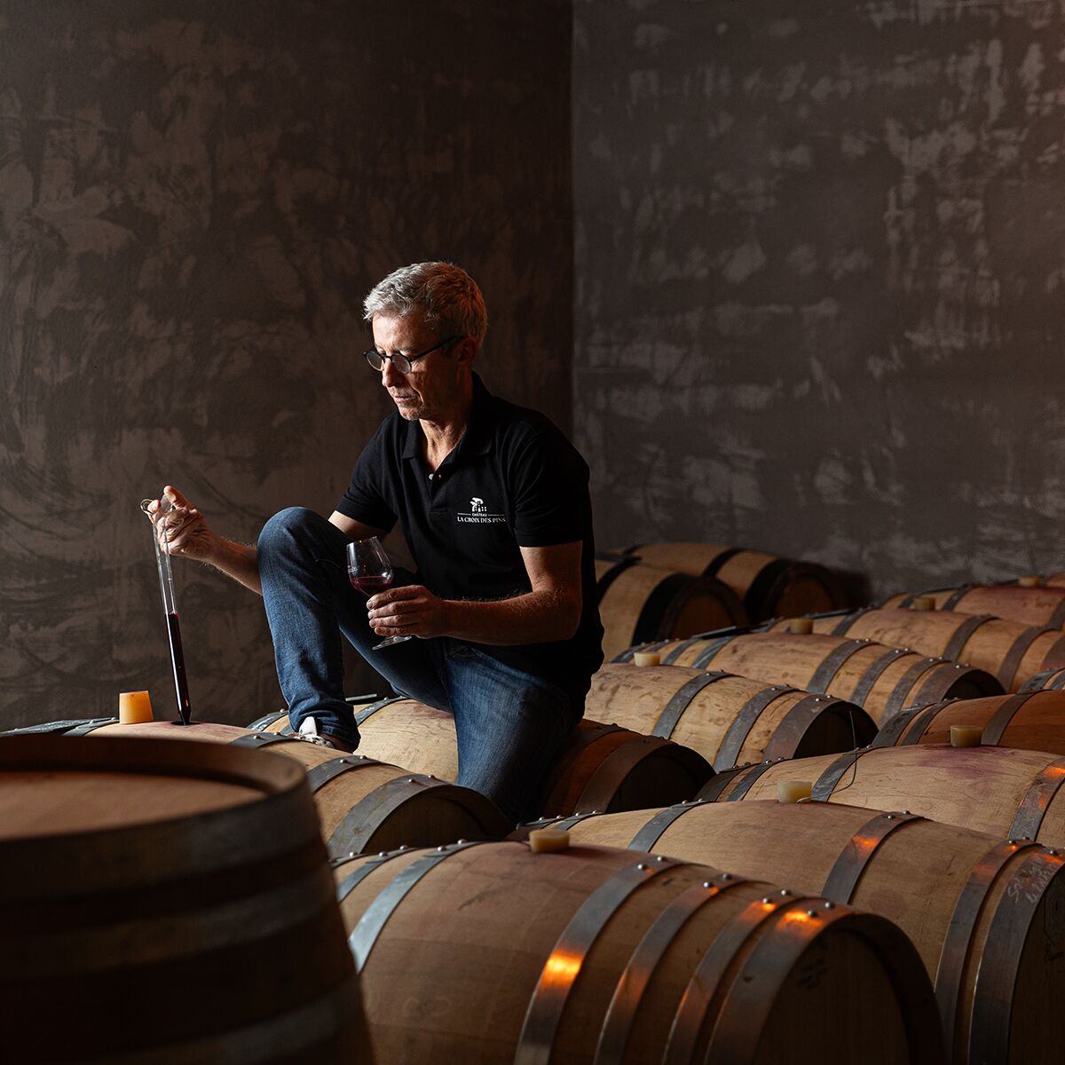 Ein Winzer sitzt in einem stimmungsvoll beleuchteten Weinkeller zwischen großen Holzfässern und prüft ein Glas Rotwein. Die Szene zeigt die traditionelle Weinherstellung und Degustation des Grenache Syrah Mourvèdre, Ventoux, La Noria aus dem 6er Paket von Croix de Pins.