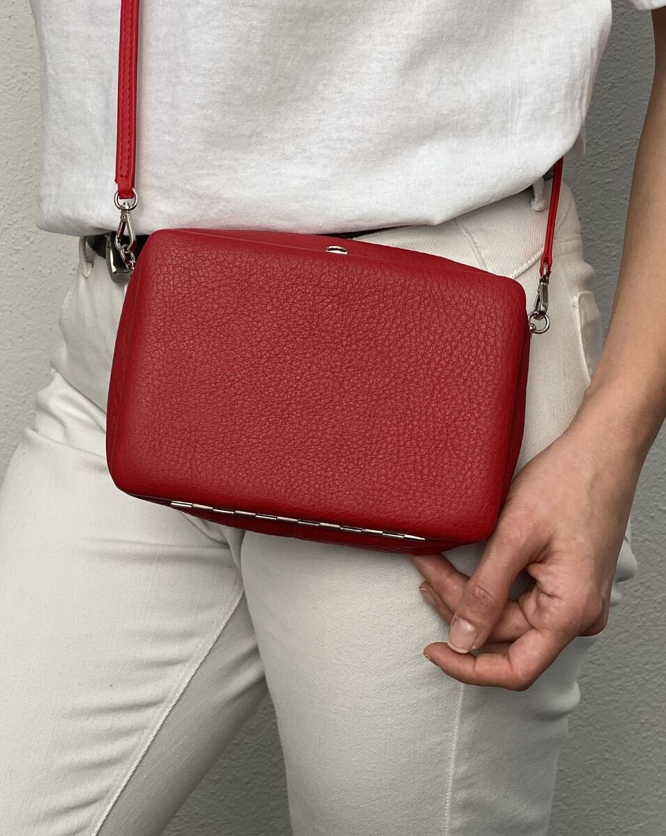 Eine rote Schultertasche aus Leder, getragen von einer Person in weißer Hose und weißem Oberteil.