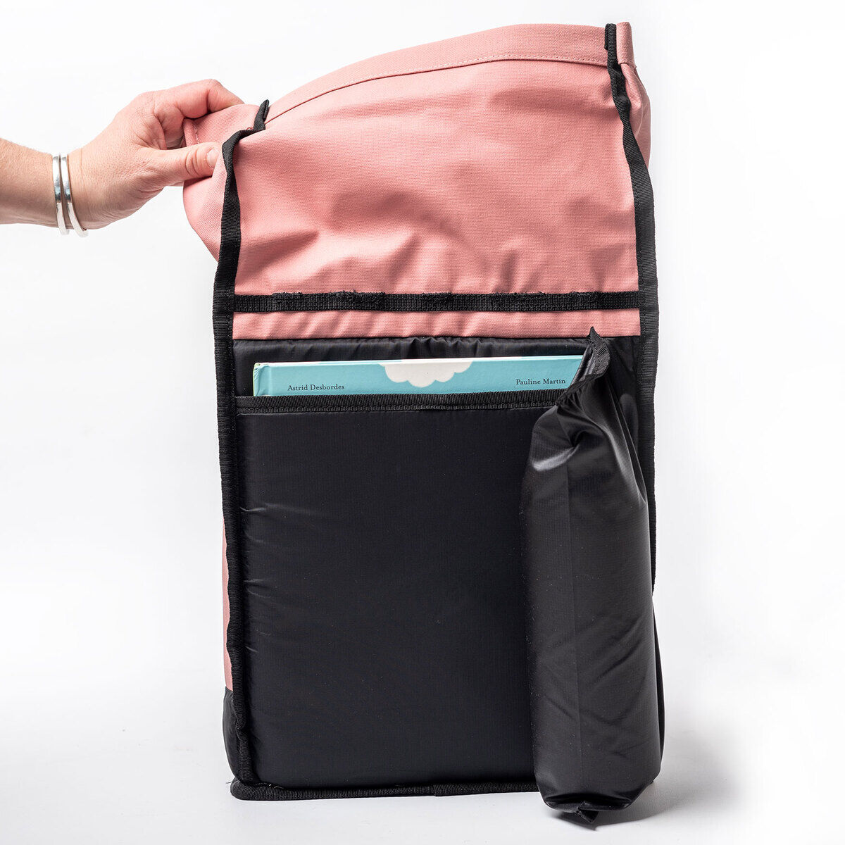 Rosa und schwarzer Rucksack „Popoyo“ auf weißem Hintergrund. Eine Hand hält den Rucksack, der halb geöffnet ist, und ein Buch ist teilweise sichtbar.