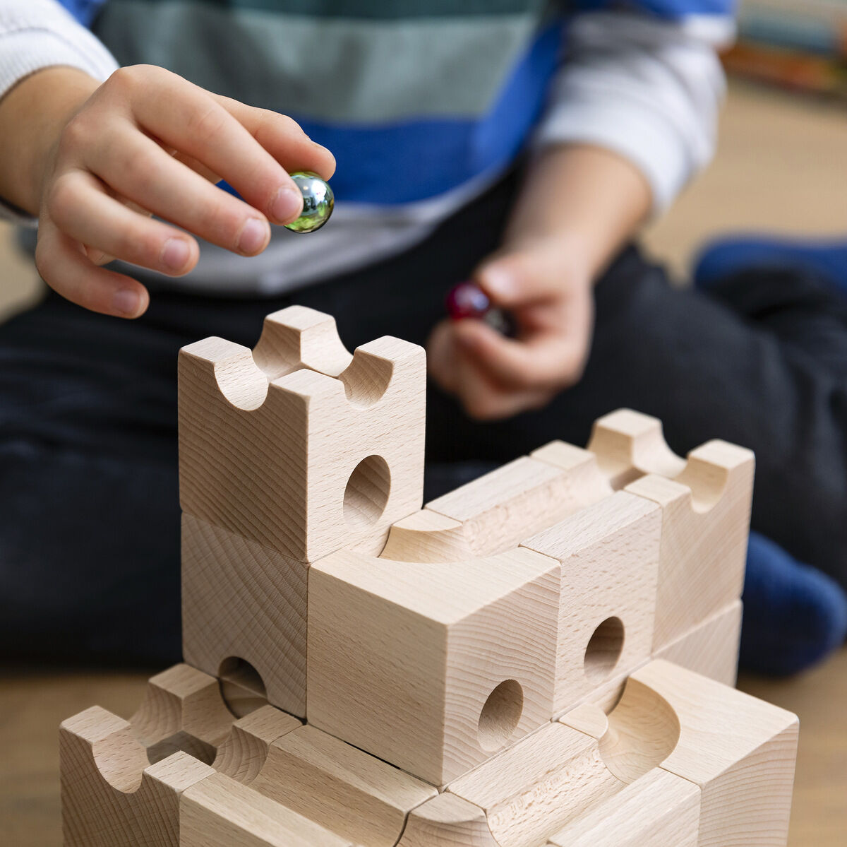 Kind spielt mit der Kugelbahn CUBORO Set »STANDARD 50 & DAS BUCH«, bestehend aus naturfarbenen Holzblöcken. Hände eines Kindes halten eine kleine Kugel über dem aufgebauten Set.