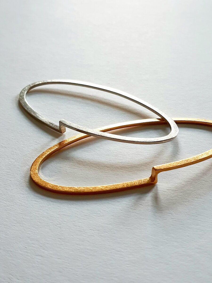 Zwei schlichte, moderne Armreifen, einer in Gold und einer in Silber, liegen überkreuzt auf einer weißen Oberfläche.