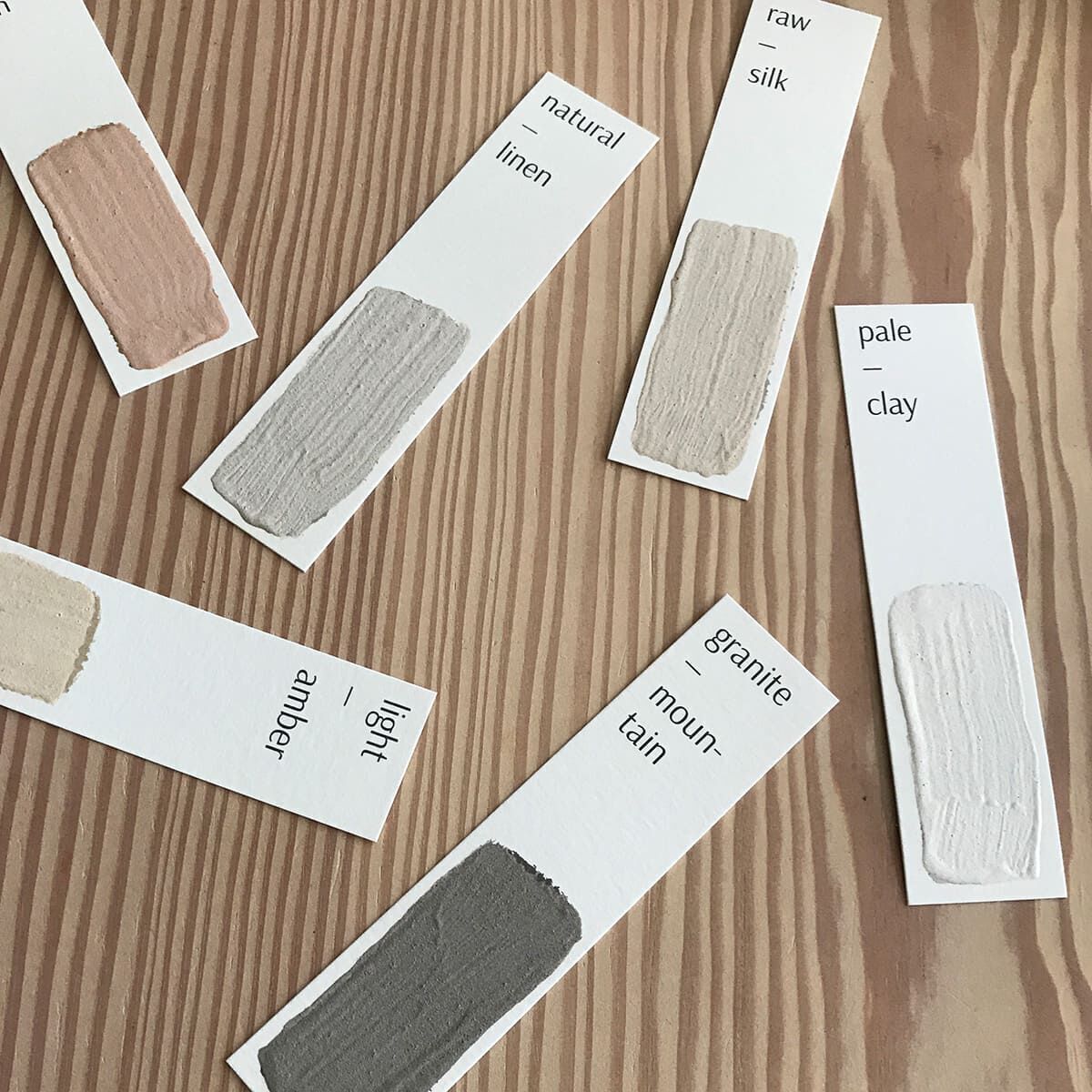 Farbfächer mit verschiedenen Lehmfarbenmustern liegen auf einer Holzoberfläche. Die Farbstreifen zeigen Töne von beige, creme, hellbraun und grau.