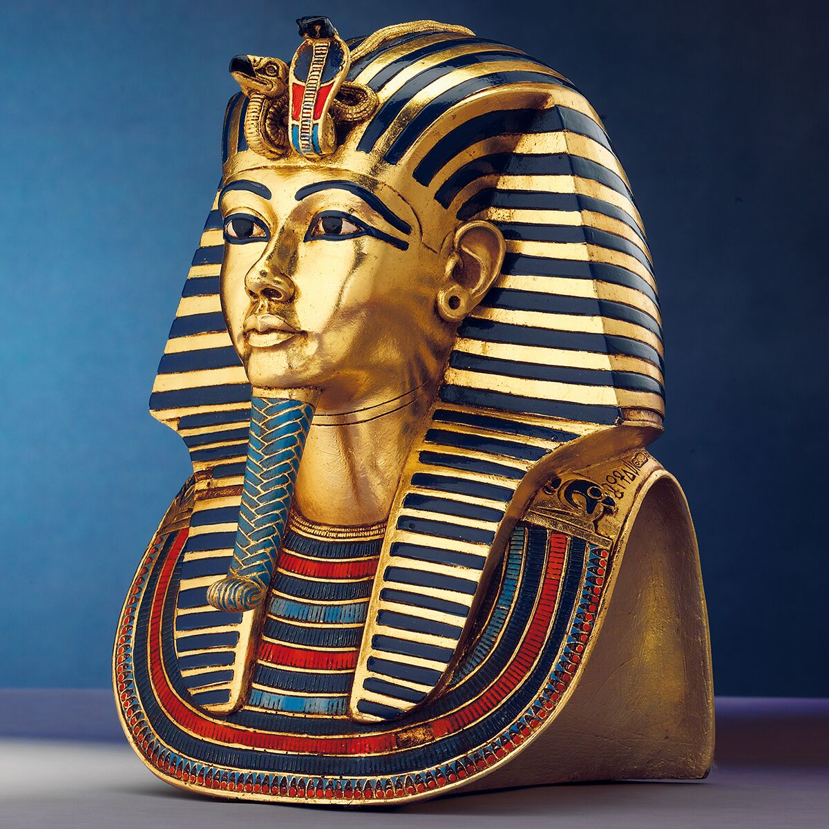 Detailreiche Büste der »Goldmaske des Tutanchamun« (Reduktion) von Ars mundi. Die Skulptur zeigt einen goldenen Pharaonenkopf mit blau-roten und schwarzen Streifen, Verzierungen und markanten Gesichtsmerkmalen vor blauem Hintergrund. Maße: ca. 24 cm hoch.