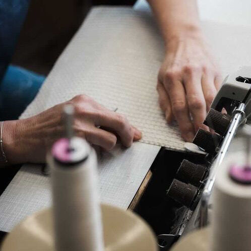 Nahaufnahme der Hände einer Person, die mit einer Nähmaschine und Werkzeugen einen Stoff bearbeitet, um ein Textilprojekt zu erstellen oder zu verändern.