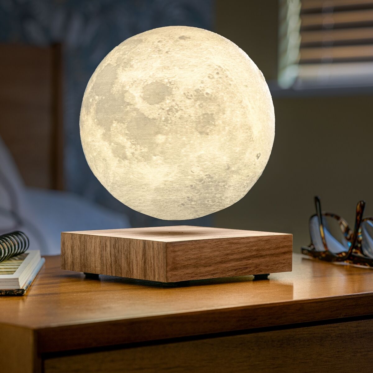 Eine schwebende Lampe in Form des Mondes auf einer Holzbasis, beleuchtet und auf einem Nachttisch stehend. Im Hintergrund sind Bücher, eine Brille und ein Bett sichtbar.