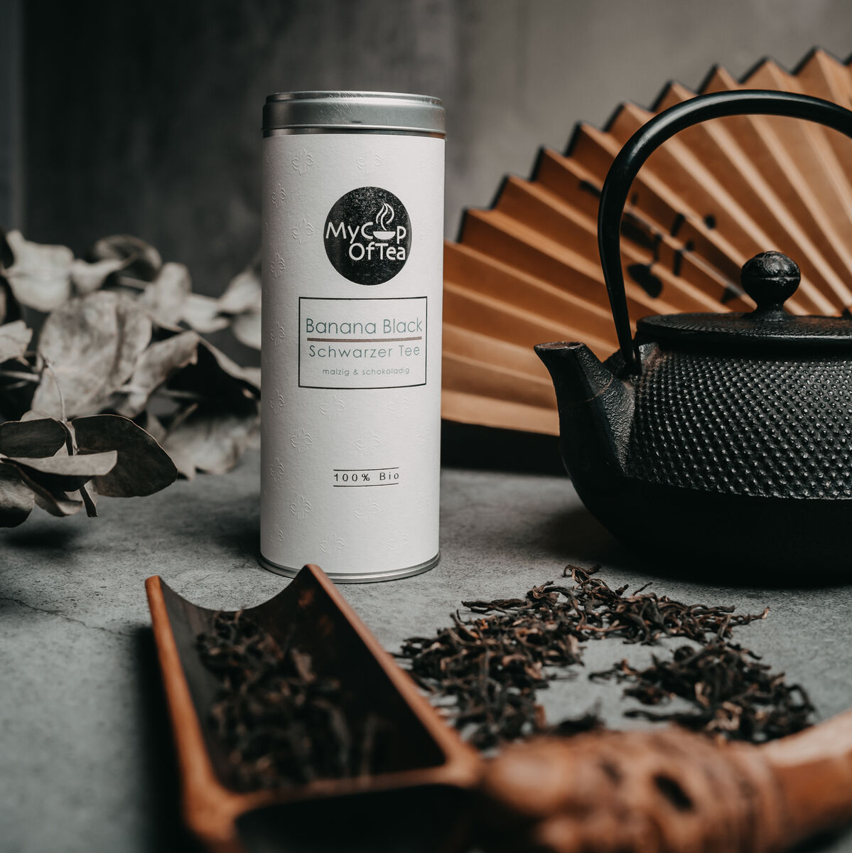 Eine Teedose von »MyOrTea« mit der Aufschrift 'Banana Black Schwarzer Tee' neben einer schwarzen Teekanne aus Gusseisen, umgeben von Teeblättern und Dekorationsartikeln auf einem grauen Untergrund.
