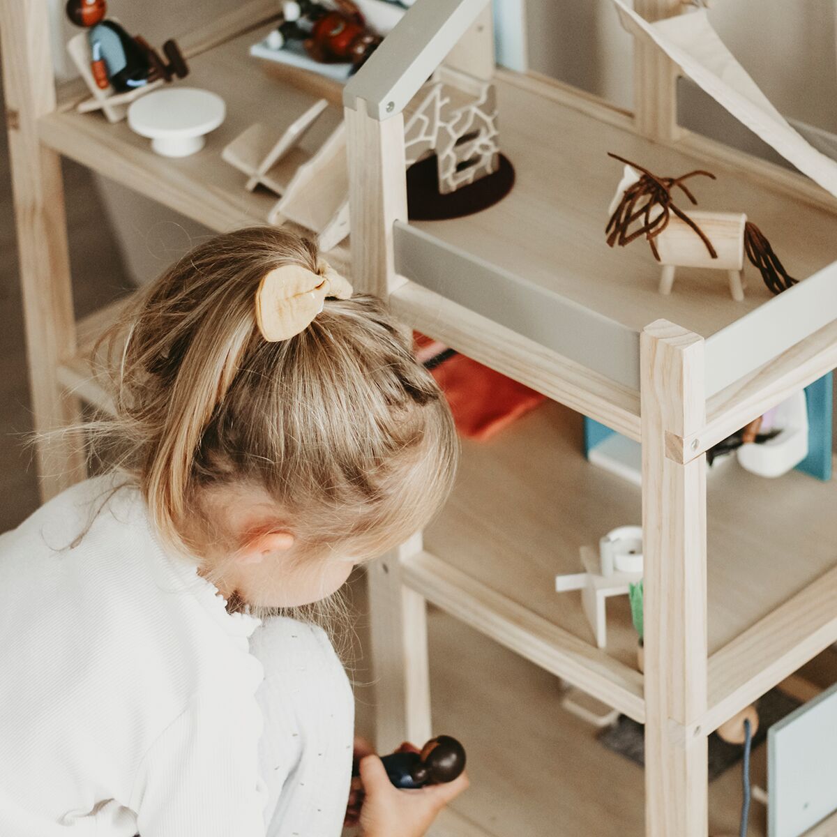 Ein Kind spielt mit einem Puppenhaus aus Holz, das mit kleinen Möbeln ausgestattet ist. Das Puppenhaus steht in einem hellen Raum auf dem Boden. Das Kind arrangiert Figuren und Möbel im Haus, das aus naturbelassenem Holz mit weißen Akzenten besteht.