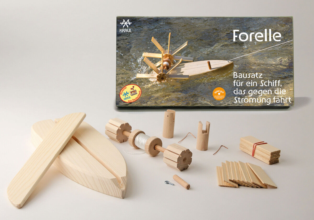 Ein Bausatz namens 'Forelle' zum Experimentieren. Enthalten sind verschiedene Holzteile wie Rümpfe, Räder, Achsen und Schnüre. Auf der Verpackung ist ein Bild eines fertiggestellten Holzschiffs, das gegen die Strömung fährt.