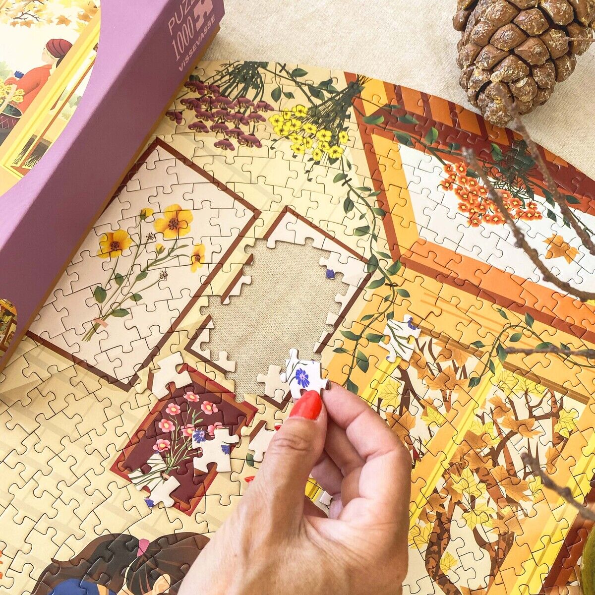Eine Hand mit rot lackierten Nägeln setzt ein Puzzleteil in ein großes, begonnenes 1000-Teile-Puzzle mit herbstlicher Szene und Blumenmotiv ein. Links liegt die Puzzle-Schachtel von VISSEVASSE, oben rechts eine braune Tannenzapfen-Deko.