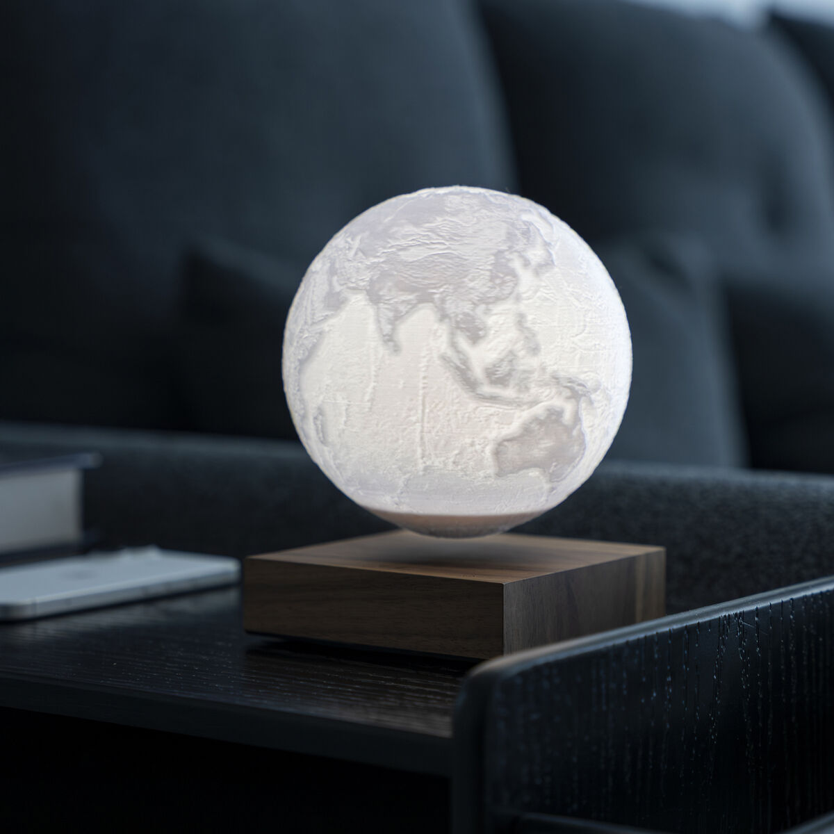 Die Leuchte »Smart Earth Lamp« von Gingko Design Ltd schwebt leuchtend über einer eckigen Holz-Basis auf einem dunklen Couchtisch. Im Hintergrund befindet sich ein modernes Sofa. Die Lampe zeigt Erdreliefs und wirkt hochwertig und dekorativ im Wohnambiente.
