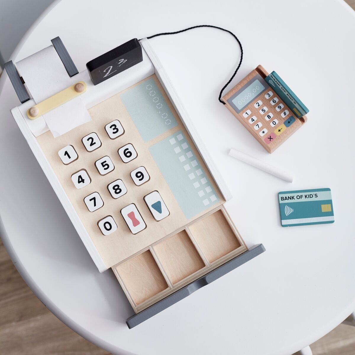 Eine Holzkasse für Kinder mit Tasten, einer kleinen Tafel, einem Kassenband und einem Spielkreditkartenleser. Das Spielzeug ist auf einem weißen Tisch platziert.