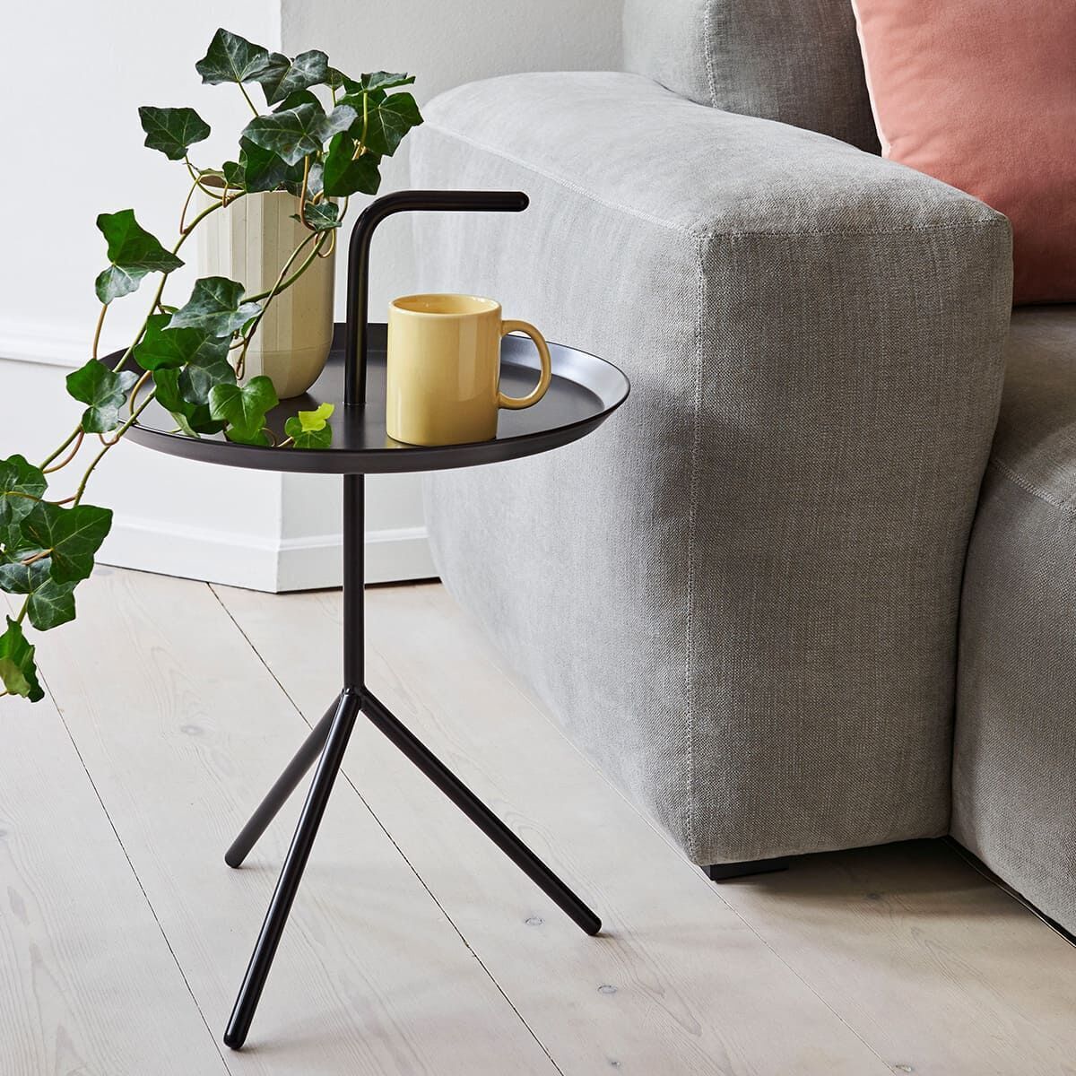Ein »DLM Beistelltisch« mit schwarzem Drahtgestell und runder Tischplatte, darauf eine gelbe Tasse, eine dekorative Pflanze und ein kleiner Keramikvasen, neben einem grauen Sofa.