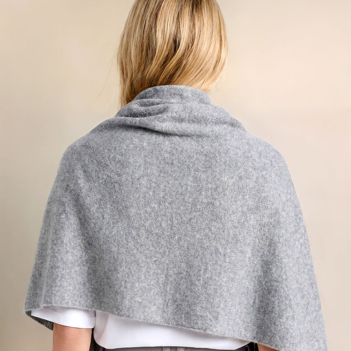 Eine Frau trägt eine graue Kaschmirstola "Donna" Graumelange lässig um Schultern und Rücken. Die Stola wirkt weich und elegant. Sie kombiniert sie mit weißem Shirt und beigefarbener Hose vor neutralem beige-cremefarbenem Hintergrund.