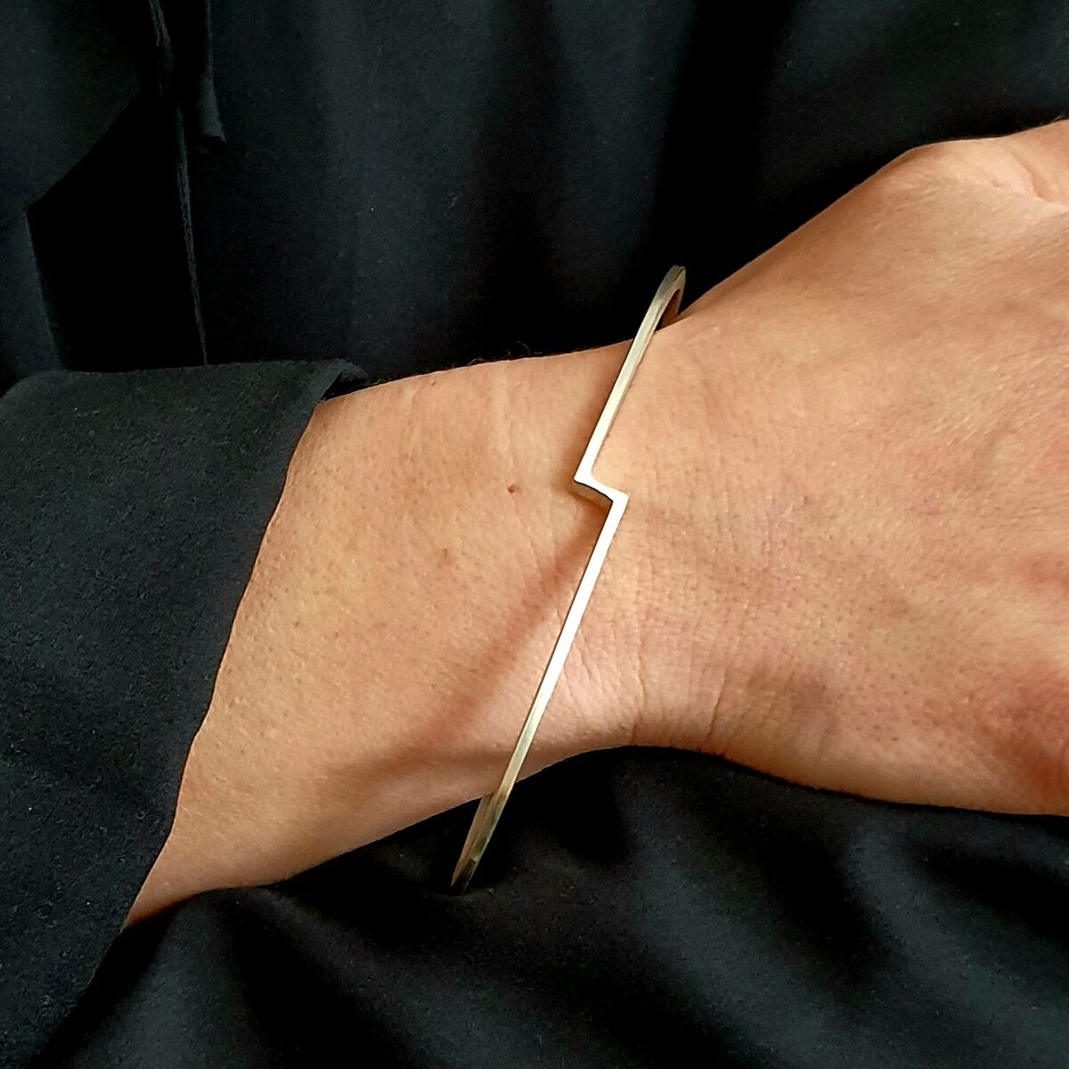 Schlanker, goldener Armreif mit einem zickzackförmigen Design, getragen am Handgelenk einer Person mit schwarzer Kleidung.