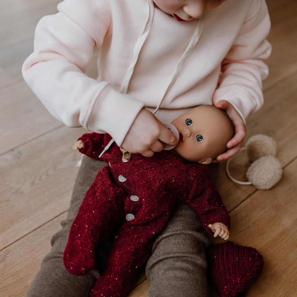 Kind sitzt auf dem Boden und spielt mit einer Babypuppe namens Muffin Baby Jubilee, die in einen bordeauxroten Einteiler gekleidet ist. Die Szene wirkt warm und einladend, das Kind steckt der Puppe einen Schnuller in den Mund. Im Hintergrund ist ein holzfarbener Fußboden zu sehen.