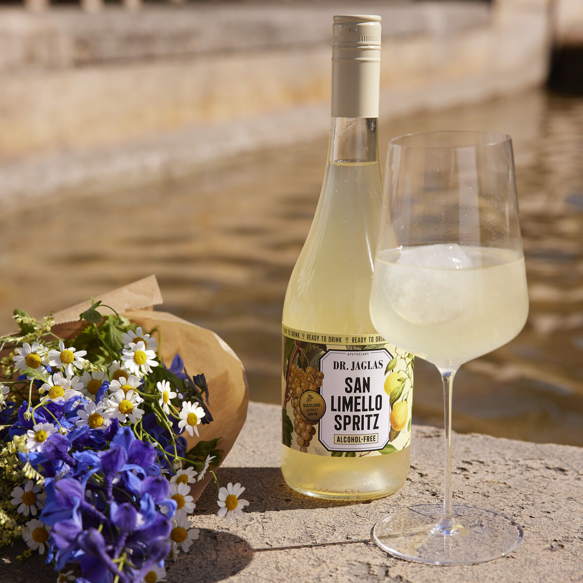 Eine Flasche San Limello Spritz und ein gefülltes Weinglas stehen auf einer Steinmauer neben einem Strauß aus blauen und weißen Blumen vor einem Gewässer.
