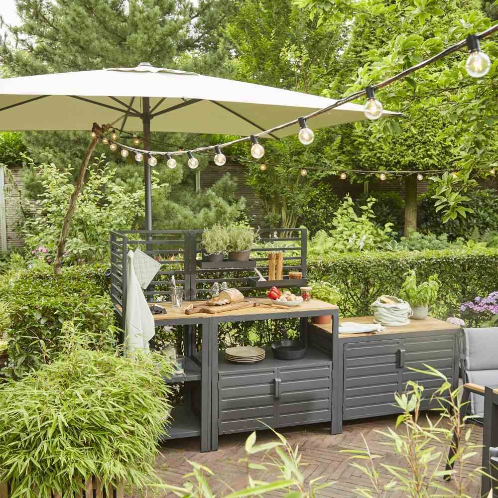 Outdoor-Küche Mittelteil »Sierra« auf einer Terrasse, umgeben von grünen Pflanzen, einem großen Sonnenschirm und einer Lichterkette, perfekt für Gartenpartys und entspannte Sommerabende.