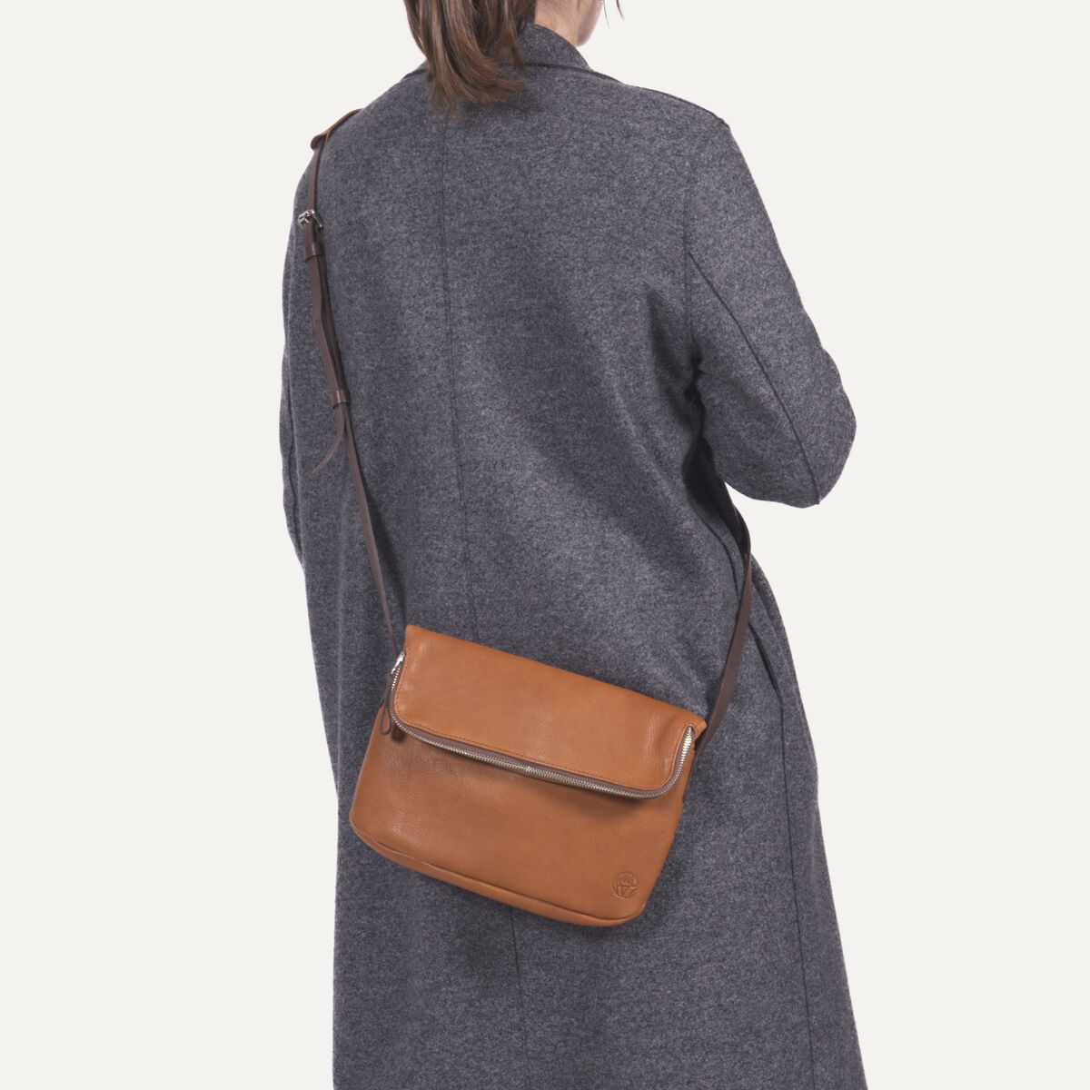 Eine Frau trägt einen grauen Mantel, helle Hose und eine braune Chaza Twoway Handtasche mit verstellbarem Schulterriemen über der Schulter. Die Tasche ist aus Leder gefertigt und kombiniert zeitloses, elegantes Design mit praktischer Funktionalität.