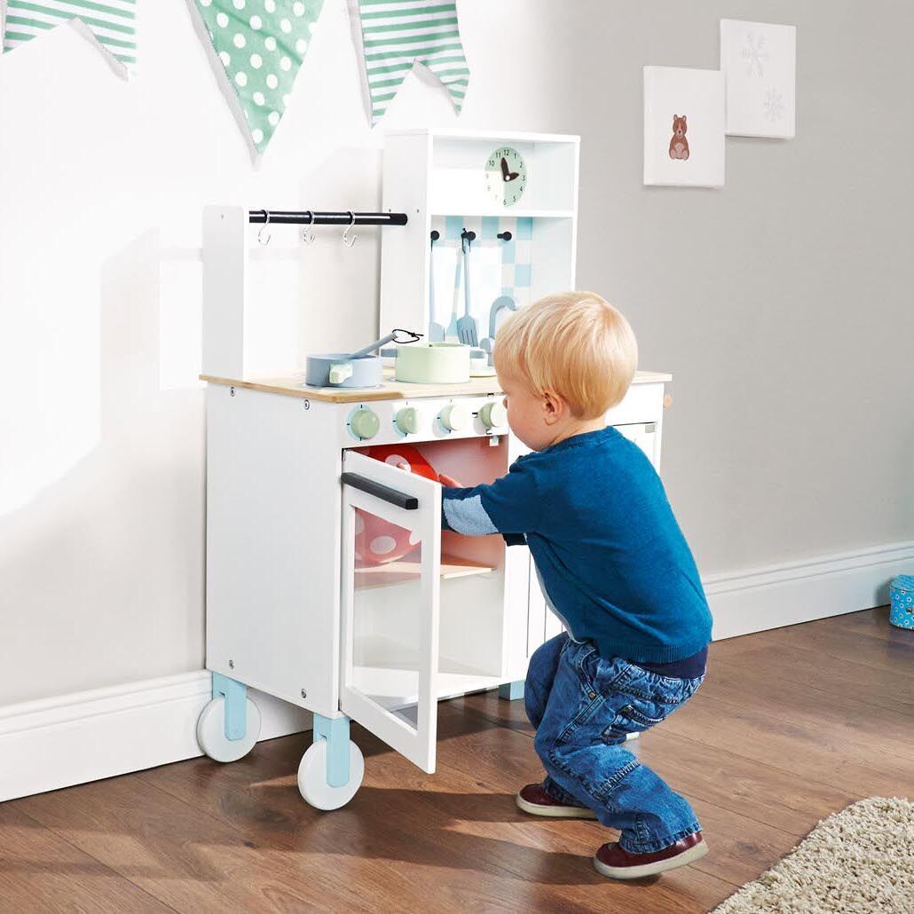 Ein kleines Kind spielt mit einer weißen Holzküche namens 'Kinder-Kombi-Küche »Alfons«'. Die Küche befindet sich in einem hellen Raum mit braunem Holzboden und an der Wand hängen grüne, gepunktete Wimpel. Die Küche hat Zubehör wie Töpfe und Pfannen und Türen zum Öffnen.