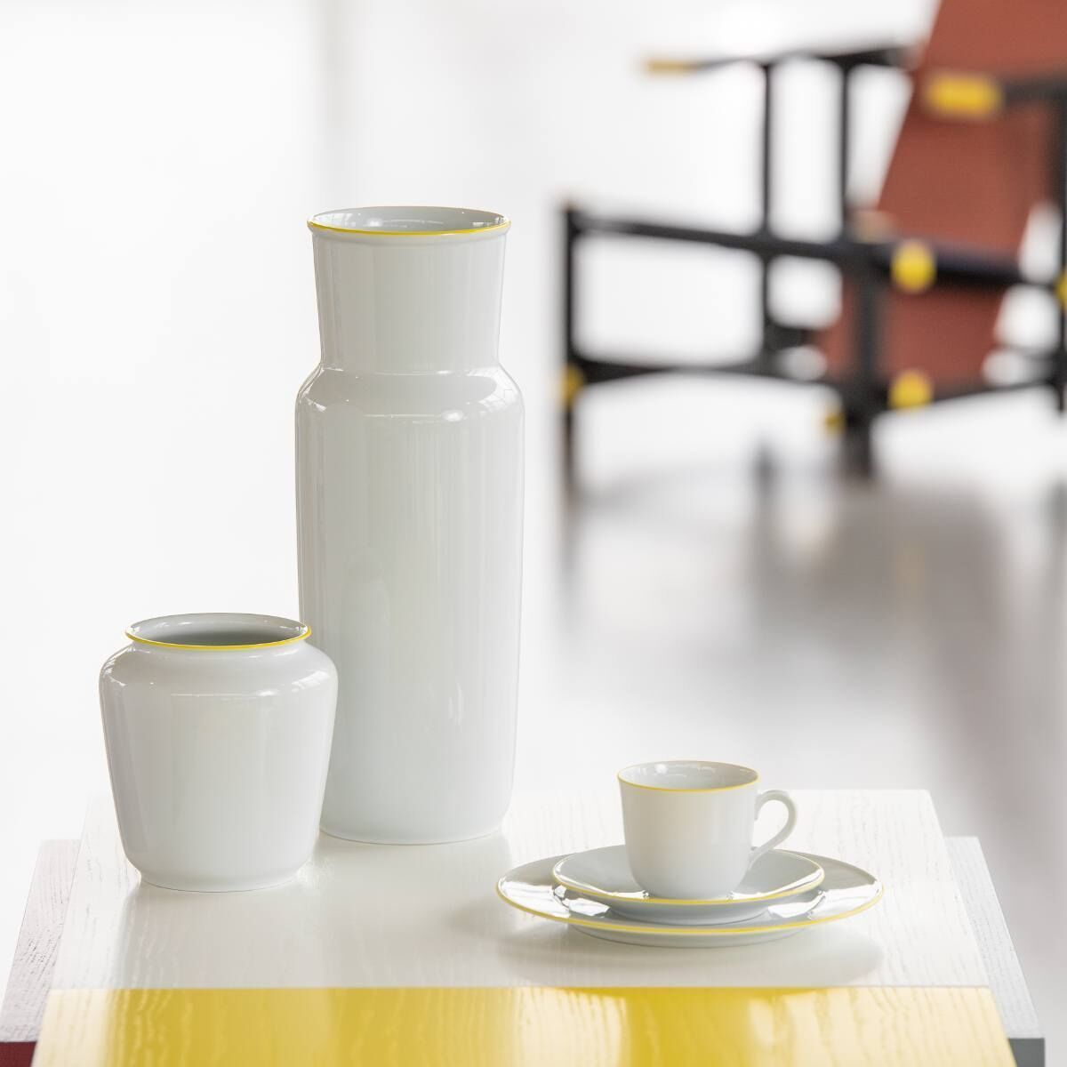 Weiße Wagenfeld Vase 639/14 von Fürstenberg, bestehend aus einer hohen und einer kleineren Vase, beide mit gelbem Rand auf einem weißen Tisch platziert. Im Hintergrund sind unscharfe Möbel zu sehen.