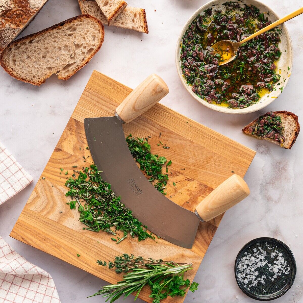 Ein Wiegemesser mit Holzgriffen liegt auf einem Holzbretthaufen, das mit frisch gehackten grünen Kräutern bedeckt ist. Daneben befinden sich Scheiben von Bauernbrot, grobes Salz in einer schwarzen Schale, eine Schüssel mit Oliven und einer Kräutermischung in Öl, sowie ein Küchentuch.