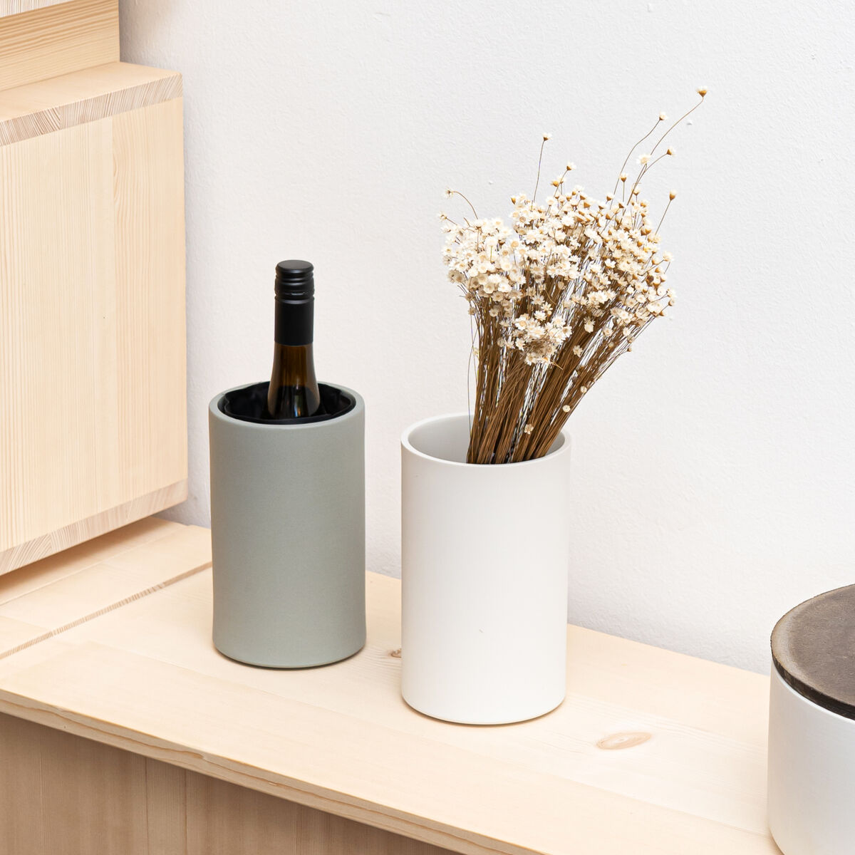 Zwei zylindrische Porzellanobjekte, einer als Weinkühler in betongrün matt mit Flasche, einer als Vase in weiß matt mit Trockenblumen. Beide stehen auf hellem Holz vor weißer Wand, rechts steht eine Dose mit braunem Deckel. Schlicht und modern.