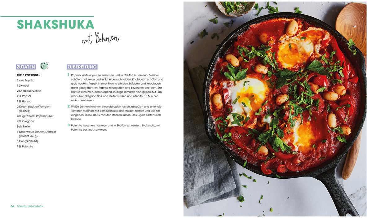 Eine Doppelseite des Kochbuchs »Studieren, kochen, leben« zeigt ein Rezept für Shakshuka. Auf der rechten Seite befindet sich ein Foto des fertigen Gerichts in einer schwarzen Pfanne mit roten Tomaten, weißen Bohnen, Eiern und grünen Kräutern. Links daneben stehen Zutaten und Zubereitung.