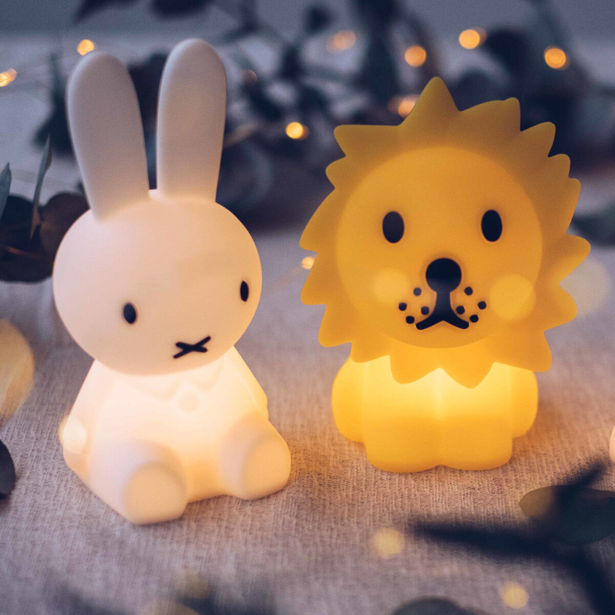 Zwei Nachtlichter in Form von Tieren, eines ist ein Löwe und das andere ein Hase, die warmes Licht spenden und auf einem Tisch mit einer grauen Tischdecke stehen, im Hintergrund sind Lichterketten unscharf zu sehen.