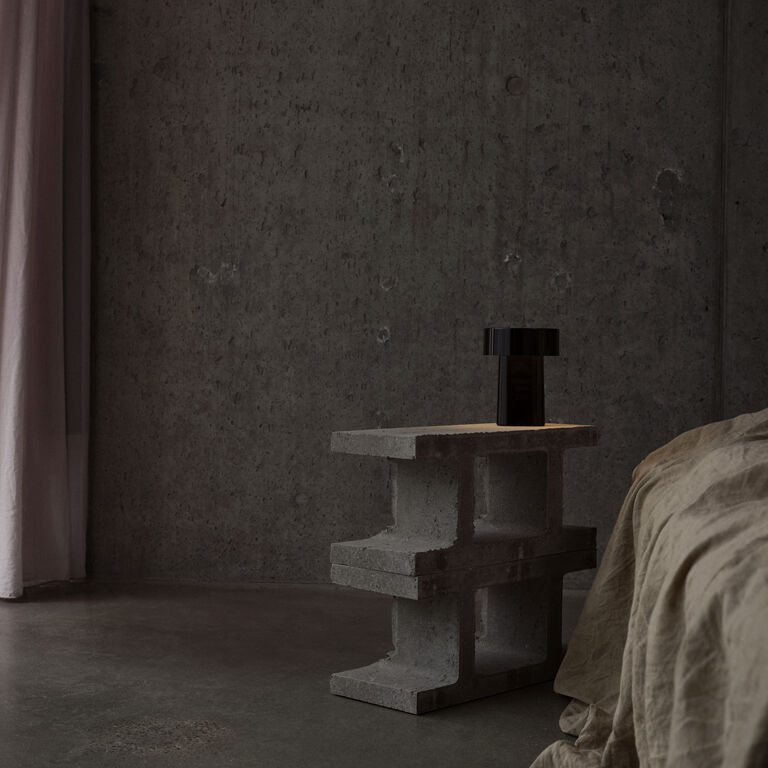 Auf einem modernen, grauen Betontisch steht eine tragbare, schwarze Lampe im minimalistischen Design. Der Raum ist dezent beleuchtet, die Wände sind aus rauem Sichtbeton, ein Bett mit beiger Bettwäsche befindet sich rechts. Tageslicht fällt durch einen zartrosa Vorhang, das Ambiente wirkt ruhig und stilvoll.