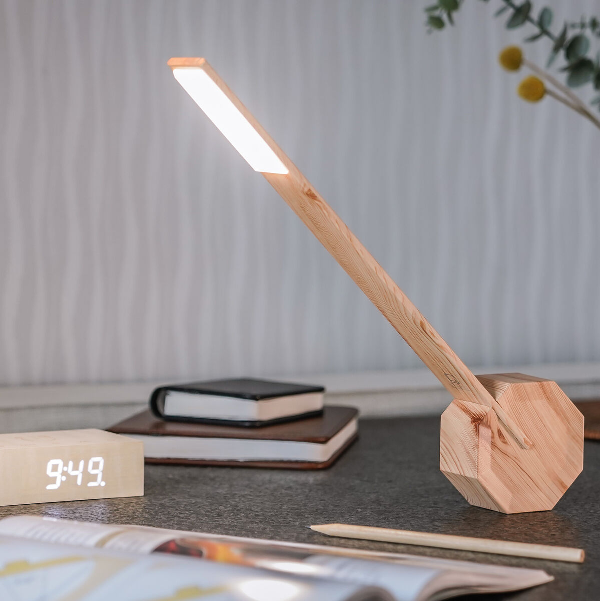 Eine moderne, minimalistische Tischlampe aus Holz mit achteckiger Basis. Die Lampe steht auf einem dunklen Tisch neben einem digitalen Wecker, einem Buch und einem Stift. Im Hintergrund ist eine Vase mit Zweigen und Blättern zu sehen.