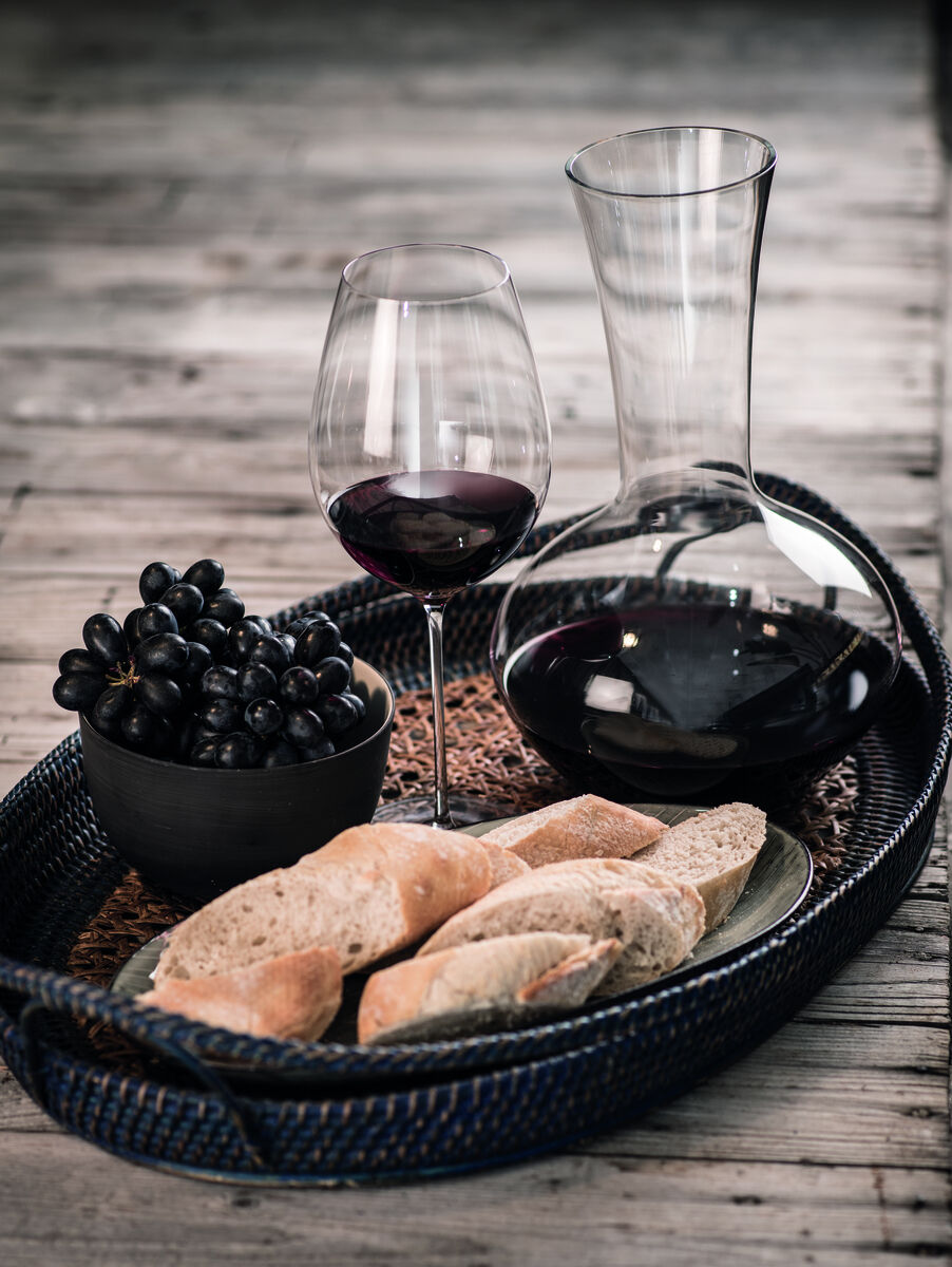 Eine Flasche Rotwein und ein Weinglas mit rotem Wein neben schwarzen Trauben in einer Schale und geschnittenem Baguette auf einem Tablett.