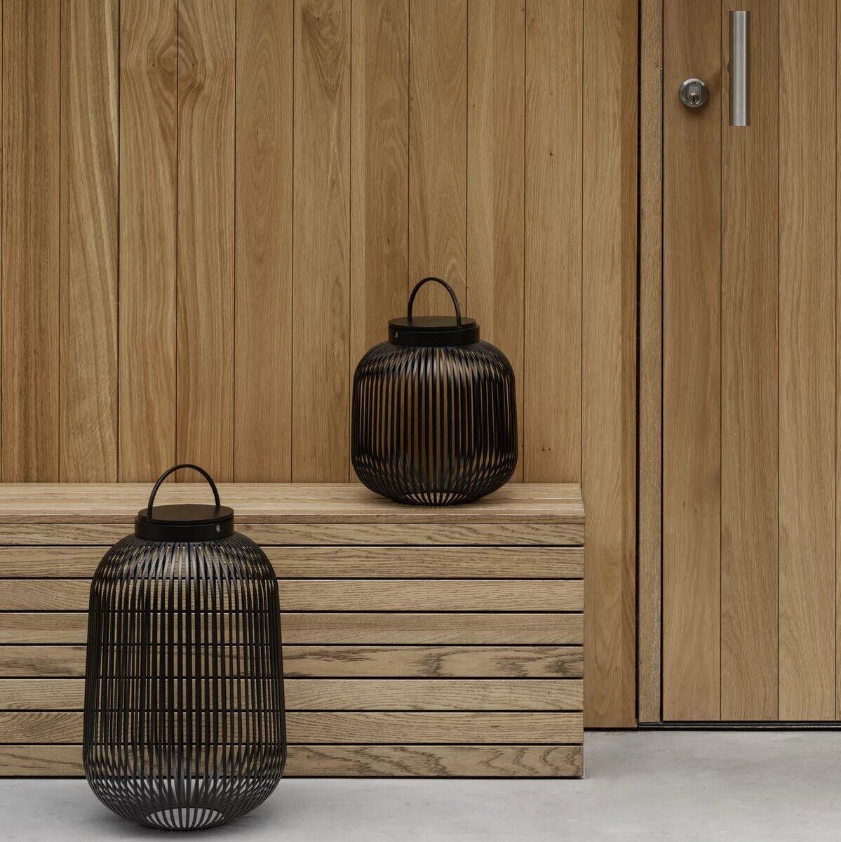 Zwei mobile LED-Leuchten der Marke LITO, Größe L, stehen auf einer Holzbank vor einer Holzwand. Die Leuchten sind schwarz und haben ein modernes, minimalistisches Design.